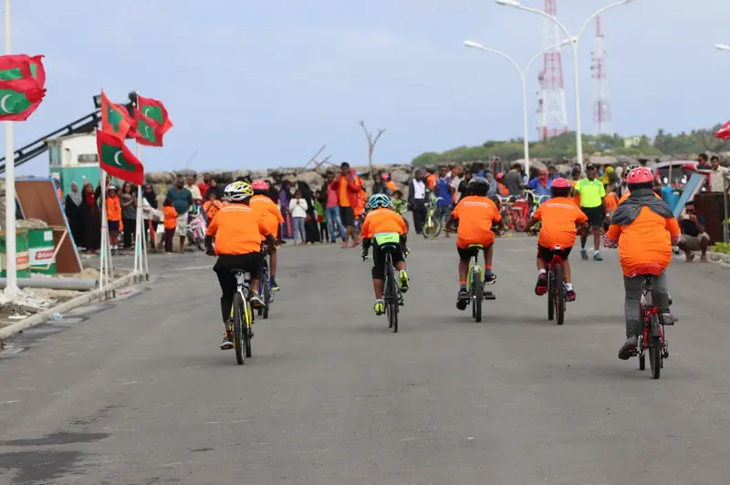 Riders in orange shirts on a seafront road with Maldivian flags